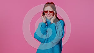 Sincere cool cheerful redhead girl in blue sweater wearing sunglasses, charming smile on pink wall