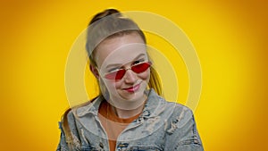 Sincere cool cheerful stylish girl in denim jacket wearing sunglasses, charming smile on yellow wall