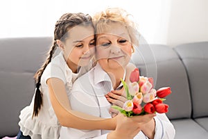 Sincere cute little granddaughter strong cuddling and kissing in cheek her 60s grandmother gave her pretty spring