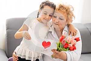 Sincere cute little granddaughter strong cuddling and kissing in cheek her 60s grandmother gave her pretty spring