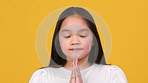 Sincere kids faith. Cute little asian girl praying to God, making prayer gesture with closed eyes and looking at camera
