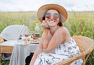 Sincerely smiling at camera young Woman in sunglasses and  straw hat dressed light summer dress sitting in rattan chair on the