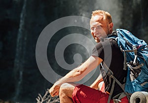 Sincerely smiling Man portrait with backpack dressed in active trekking clothes sitting near mountain river waterfall smiling and
