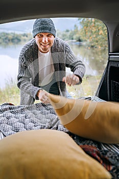 Sincerely smiling middle-aged man dressed in warm knitted clothes preparing a sleeping place in cozy car trunk on beautiful