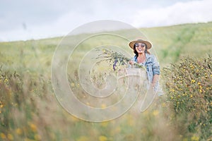 Sincerely smiling young Woman dressed jeans jacket and light summer dress walking by the high green grass meadow with basket and