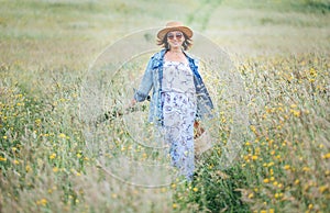 Sincerely smiling young Woman dressed jeans jacket and light summer dress walking by the high green grass meadow with basket and photo