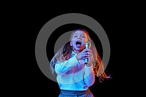 Singing with microphone. Emotional cute little girl, kid posing isolated on dark studio background in neon light