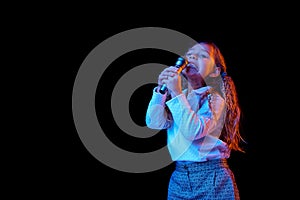 Singing with microphone. Emotional cute little girl, kid posing isolated on dark studio background in neon light