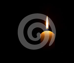 Single burning candle flame or light glowing on a big white candle on black or dark background on table in church for Christmas,