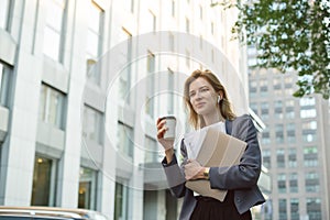 Single calm smiling businesswoman with folded hands walking the street with coffee, documents and laptop. Cheerful young female