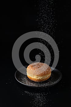 Single fresh baked donut sprinkled with falling sugar powder on dark retro plate on black background. Berliner donut