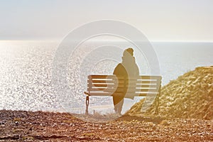 Single girl in a black jacket and hat sitting on bench at cliff at front of sea peaceful quiet place