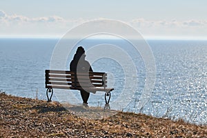 Single girl in a black jacket and hat sitting on bench at cliff at front of sea peaceful quiet place