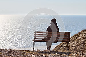 Single girl in a black jacket and hat sitting on bench at cliff at front of sea peaceful quiet place