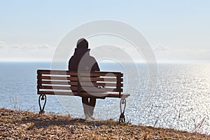 Single girl in a black jacket and hat sitting on bench at cliff at front of sea peaceful quiet place