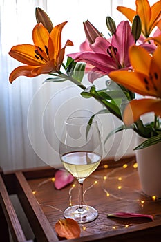 Single glass of white wine, a bouquet of lily flowers and festive lights on a wooden table table