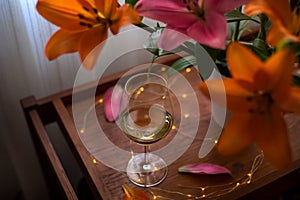 Single glass of white wine, a bouquet of lily flowers and festive lights on a wooden table table. Still life romantic evening