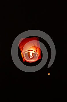 A single glowing orange sky lantern rises against a dark night background, surrounded by faint lights of other lanterns