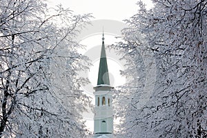 Single green and white color minaret of a small mosque in winter. Trees with rime and snow around.