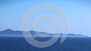 Single lonely sailing boat in sea against misty island and windmill farm slhouette - panning