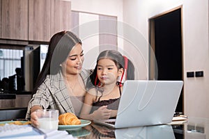 a single mother, sits at home working on a laptop with her daughter beside her watching and encouraging her.