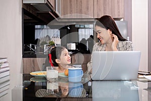 a single mother, sits at home working on a laptop with her daughter beside her watching and encouraging her.