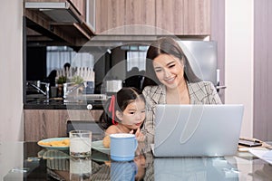 a single mother, sits at home working on a laptop with her daughter beside her watching and encouraging her.