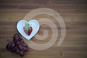 Food photography image of healthy strawberry in love heart shape dish with purple grapes and rustic wooden background