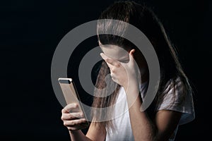 Single sad teen holding a mobile phone lamenting sitting on the bed in her bedroom with a dark light in the background