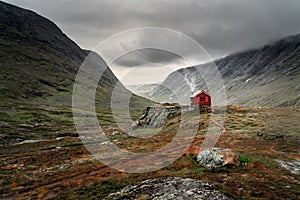 A single small red cabin billows smoke from it`s tiny chimney in the remote gorge of central Norway. The distant dark clouds give