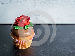 Single vanilla cupcake with chocolate frosting decorated with a red rose and green leaves against a black slate and gray shiplap