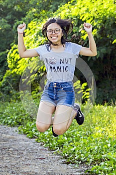 Single young trendy energetic Asian lady with long hair spectacles wearing short denim jean jump for joy at park.