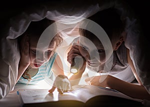 Sister and brother are reading a book under a blanket with flashlight. Pretty young boy and lovely girl having fun in children roo