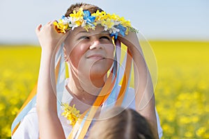 Sister puts on brother Ukrainian wreath with ribbons, against background of fields and sky