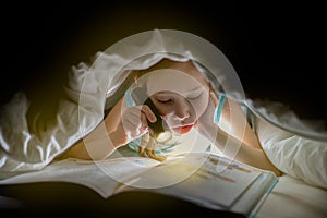 Sister are reading a book under a blanket with flashlight. Pretty young girl having fun in children room.