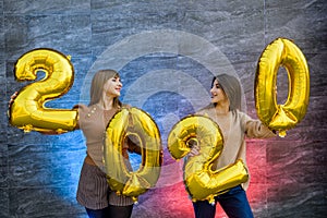 Sisters celebrating new 2020 year. They holding golden air balloons at party celebration