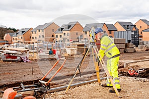 Site engineer in hi-viz working on house building construction site using modern surveying equipment against new houses