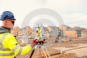 Site engineer in hi-viz working on house building construction site using modern surveying equipment against new houses nad blue