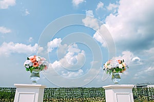 Site for the wedding ceremony outdoor. Fresh flowers in glass vases stand on white tables against blue sky with white clouds