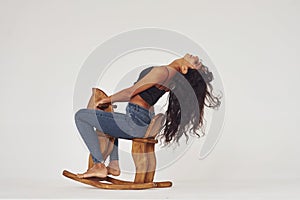 Sits on kid`s wooden horse toy. Young woman in jeans and bra indoors. White background