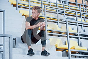 Sits on stairs. Sportive young guy in black shirt and pants outdoors at daytime