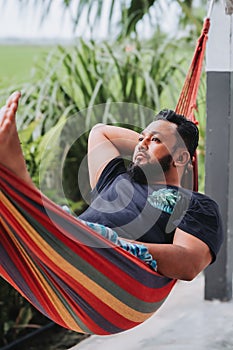 Sitting in a cozy hammock in a verdant garden close to the paddy field is a bearded Asian man.