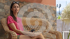 Sitting girl looking at camera smiling, holding device in a sofa. Young woman relaxing in a lounge couch at the terrace. Teenager