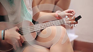 Sitting girl in shorts tune brown ukulele guitar on street. Summer sunny day. Music. Strings. Sound