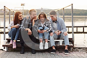 Sitting near lake and embracing each other. Group of young cheerful friends that is outdoors having fun together