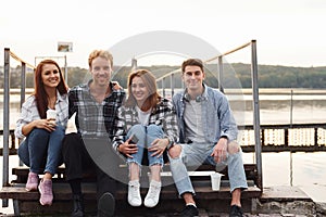 Sitting near lake and embracing each other. Group of young cheerful friends that is outdoors having fun together