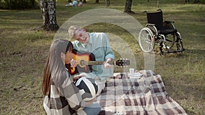 Sitting on the plaid in the public park, a disabled young man offers his girlfriend playing guitar and singing songs