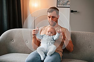 Sitting on the sofa. Father with his newborn baby is indoors. Conception of single dad