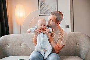 Sitting on the sofa. Father with his newborn baby is indoors. Conception of single dad