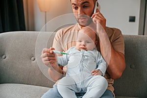 Sitting and talking by phone. Father with his newborn baby is indoors. Conception of single dad